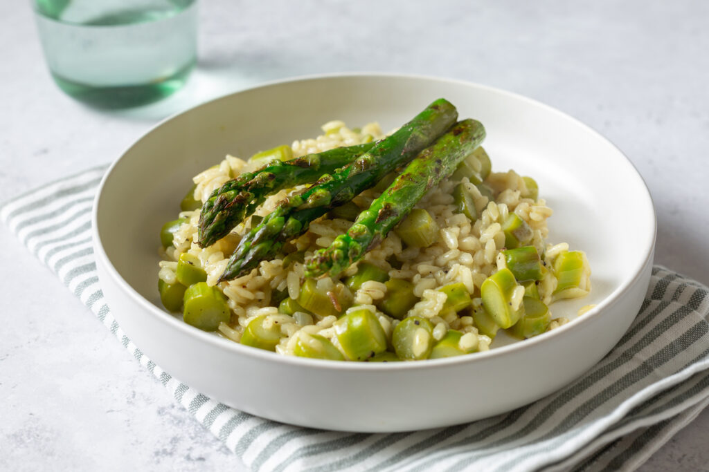 Delicious risotto with grilled asparagus on white plate, grey background with green glass, horizontal