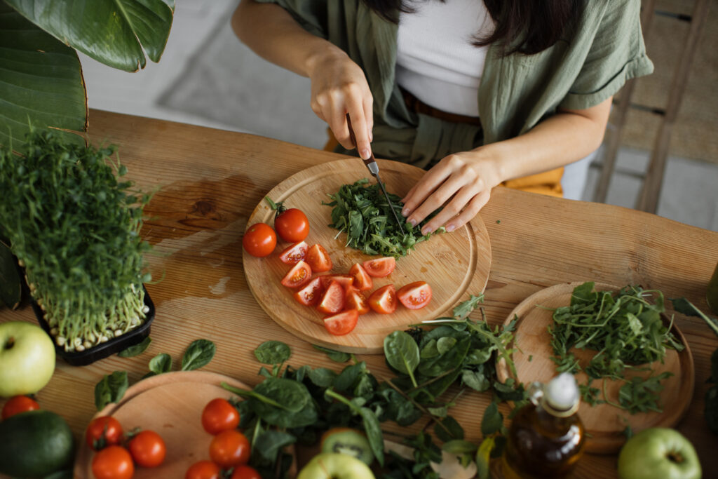 High angle view of hands of young woman chopping arugula leaves, sitting at table in bright exotic studio with many fresh fruits and vegetables are preparing for cooking.