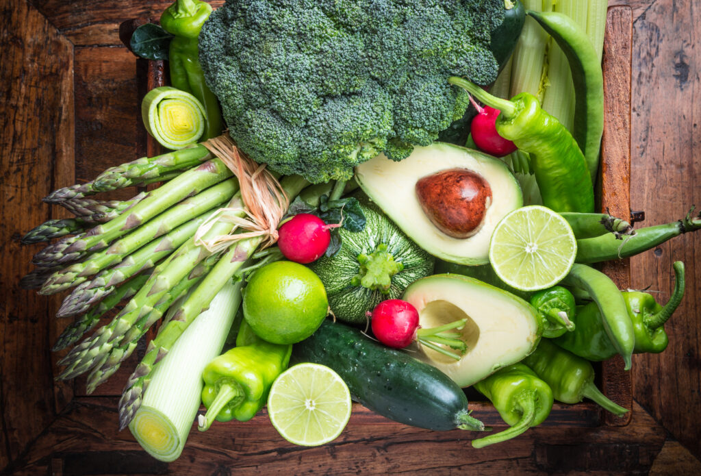 Mix of fresh green vegetables on rustic wood background. Peppers, cabbage, celery, avocado, asparagus, cucumber, radishes, broccoli, limes, beans, peas, leek.